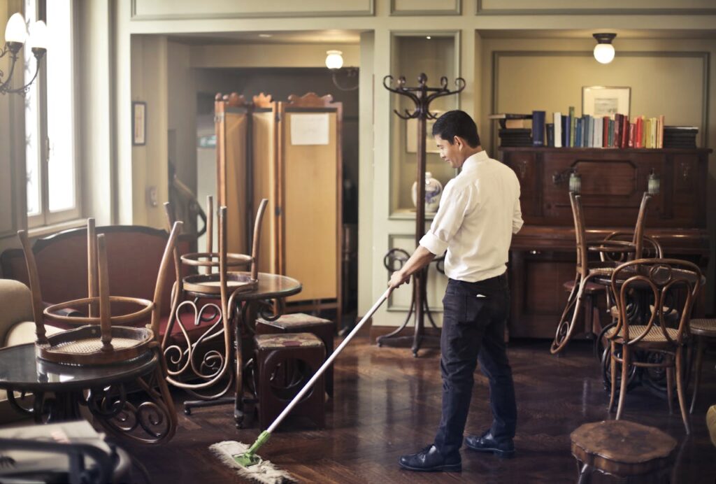 A man mopping the floor in a vintage style café with stacked chairs and warm lighting.