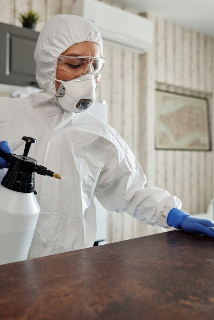 Person in protective suit disinfecting indoor surfaces with spray.