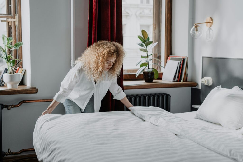 A woman with curly hair making a bed in a bright, cozy bedroom with plants and natural light.