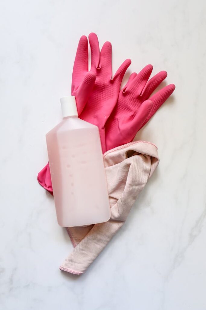 A flat lay of pink rubber gloves, a cleaning cloth, and a bottle on a white background for cleaning themes.