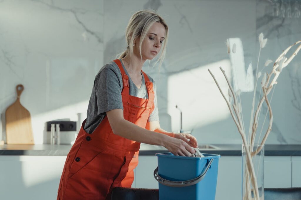 A woman in a gray shirt and red overalls cleans a kitchen counter using a blue bucket and white rag. Modern setting.