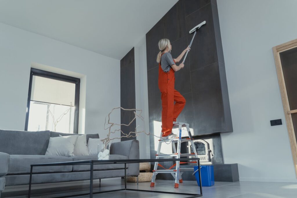 A woman stands on a ladder in a modern living room, cleaning the high wall with a mop.