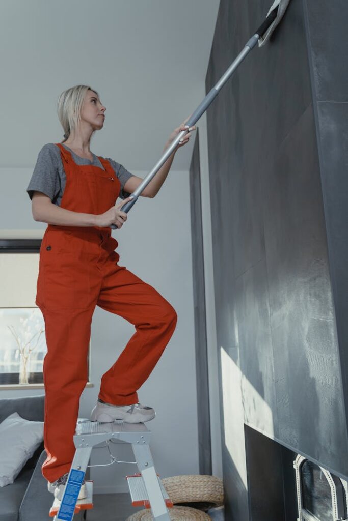 Woman in red overalls cleaning a wall using a mop while on a ladder indoors.