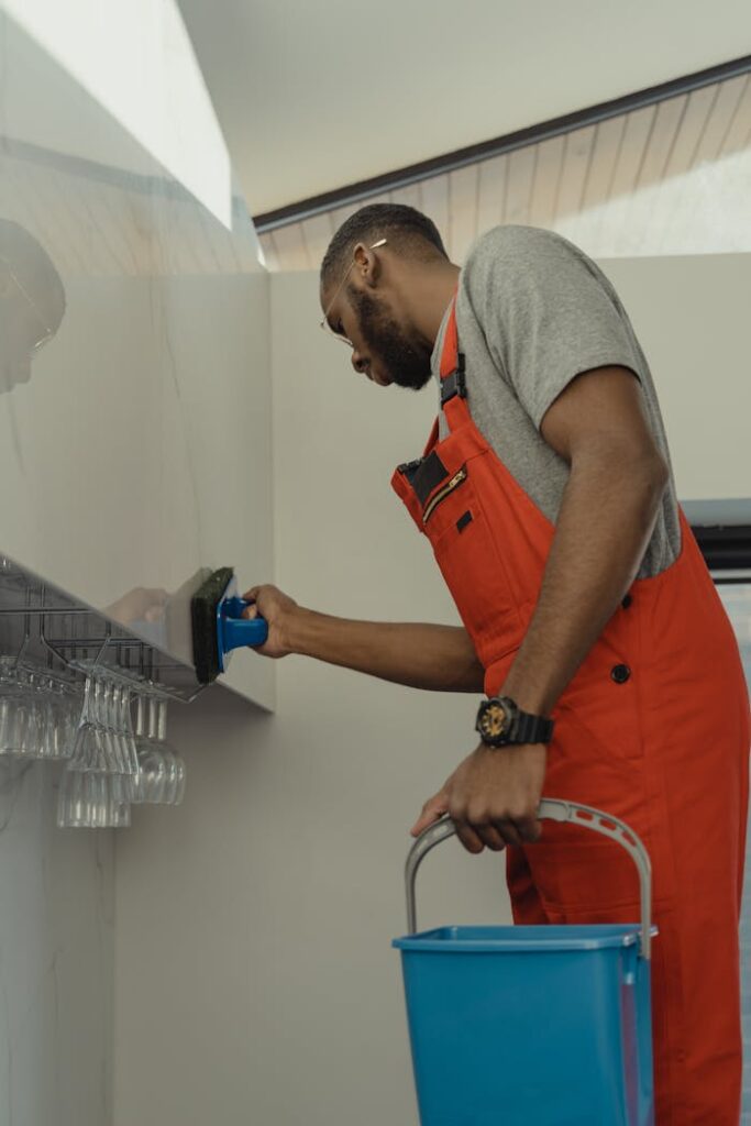 Man in red overalls cleaning a glass wall indoors with a blue bucket and brush.