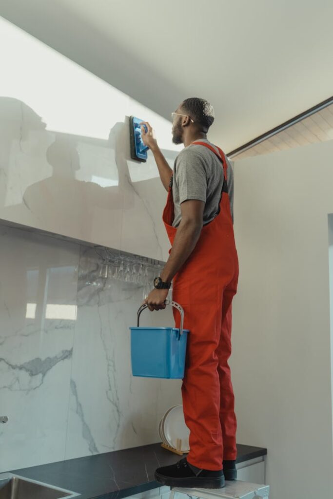 African American man in red overalls cleaning a kitchen wall with a brush and pail.