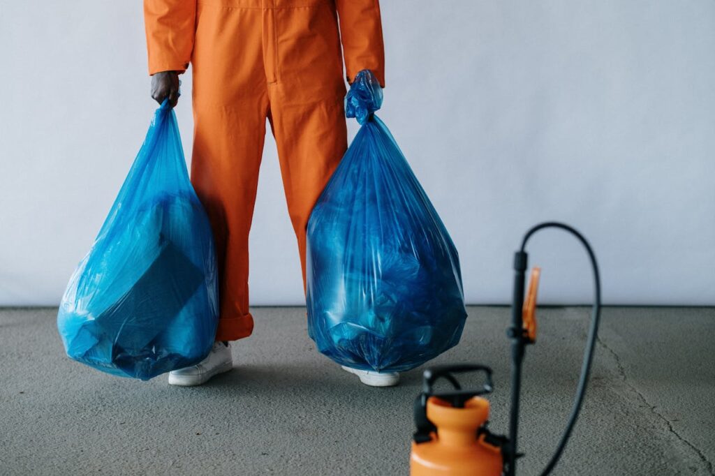 Sanitation worker in orange overalls holding blue trash bags indoors for waste management.