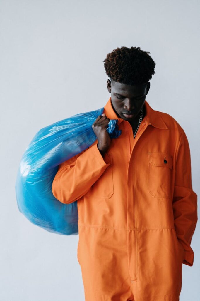 Young man in orange jumpsuit holding a blue trash bag, looking down thoughtfully in front of a white background.